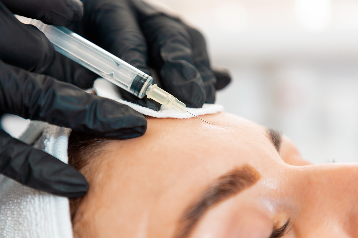 Close-up of female’s head getting injection at her forehead in the cosmetology salon. Cosmetologist in latex gloves with syringe injects a medicine. Concept of plastic surgery and face lifting